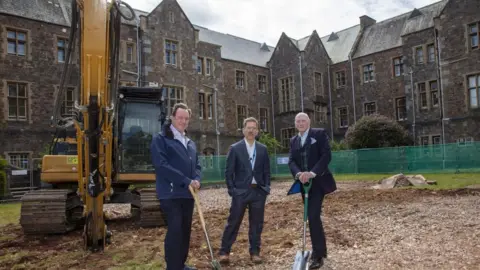 Left to right: Graham Rich, chair of Vanguard Healthcare Solutions, Tobit Emmens, associate director of research and development at DPT and Andy Willis, chair of DPT. The three men are stood on a construction site in suits. The two men on either side are holding shovels. Behind them is a yellow digger. Behind that is green fencing and a grey building. The sky is grey and cloudy. 