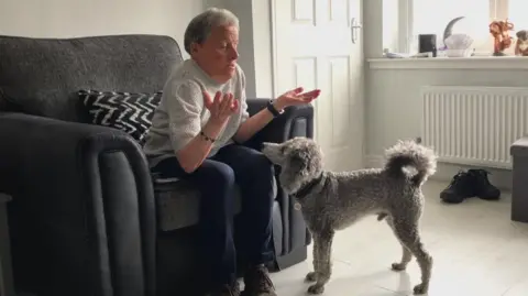 BBC/Jack Hadaway-Weller A woman sits in a living room chair with both hands raised, facing a curly-haired dog standing in front. The dog looks up attentively, positioned near the person’s knees as if awaiting instruction or responding to a cue.