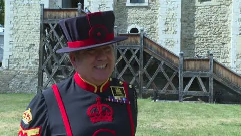 Close-up of smiling man wearing a black-and-red ceremonial uniform and hat standing outside in front of a stone castle wall and wooden staircase.