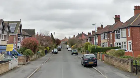 Google A street view from the centre of the road in Costock Avenue. There are rows of houses either side with cars parked on both sides of the road. 