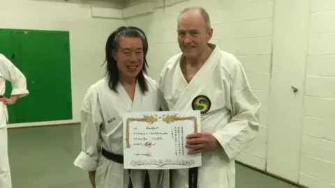 Supplied Two people wearing white martial arts uniforms with black belts stand on a padded training floor, holding a framed certificate between them. The setting appears to be an indoor dojo or training room with white brick walls, exposed yellow and white pipes along the ceiling, fluorescent lighting, and a green door in the background.