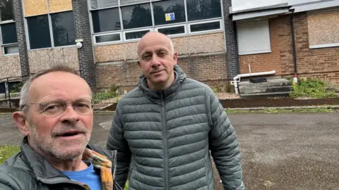 Darren Padgett Two men are stood outside a derelic building. One is balding with a grey stubble beard and glasses and the second man is balding and wearing a grey puffer jacket