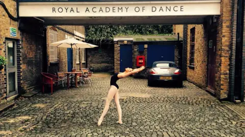 Charlotte Fastiggi A female ballet dancer poses in a cobbled street. She wears white tights, a black leotard and arches back. IN the background is a 