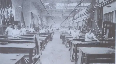 British Rail A black and white image of a large workshop with two rows of tables stretching into the distance, spaced closely together with an aisle separating them. A woman stands at every table, facing the camera. One can be seen with her hands resting on a door or window lying on her desk. The ceiling is so high that the roof cannot be seen but there are windows high up on the far wall.  