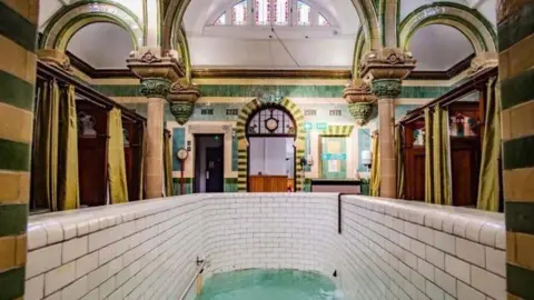 A highly decorated room with green and mustard tiles and ornate columns. There is a deep plunge pool in the foreground with white tiles. At the sides there are booths separated by wooden panels with decorations at the top and green privacy curtains.