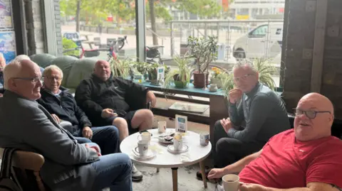 A group of five elderly men sat round a table with hot drinks. The three on the left are wearing dark colours and the one on the right is wearing red. He is bald. The other man is wearing blue.
