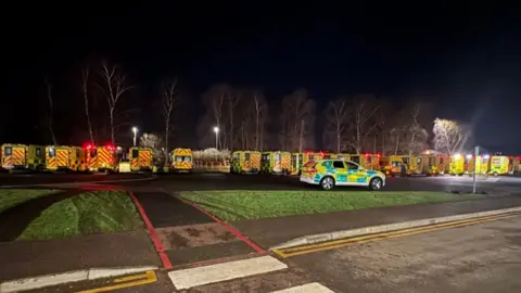 A nighttime shot of about a dozen ambulances queued up in a parking lot under lighting, with a police vehicle in the foreground