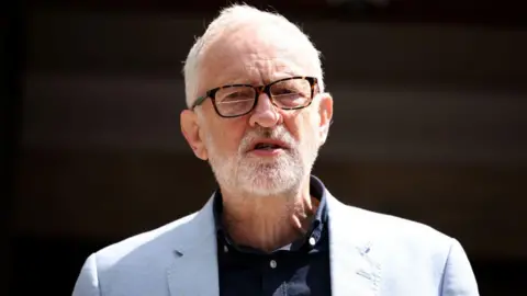 Jeremy Corbyn is seen from the shoulders up outside on a bright day wearing a light blue blazer and navy shirt.