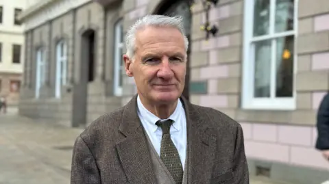 Tom Binet pictured in the Royal Square with the States building visible behind him. He's wearing a brown suit, white shirt and green tie. He has short white hair and is smiling. 