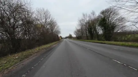A shot of the road from the perspective of a car travelling down the road. There are trees and grass at the sides of the road and a grey sky, as well as cars and a road sign in the distance. 