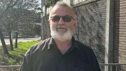 Man in black shirt with sunglasses and white beard standing outside.