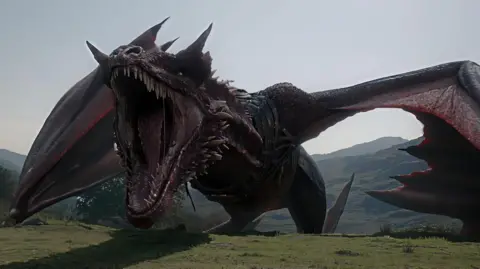 CGI of a giant red-black dragon, open mouthed, its wings out-stretched as it appears to stride rather than fly atop a mountainous landscape