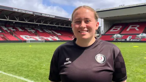 Grace, who has blonde hair pulled back into a low ponytail, smiles at the camera. She wears a black Bristol City training top, and the Ashton Gate pitch can be seen clearly behind her.
