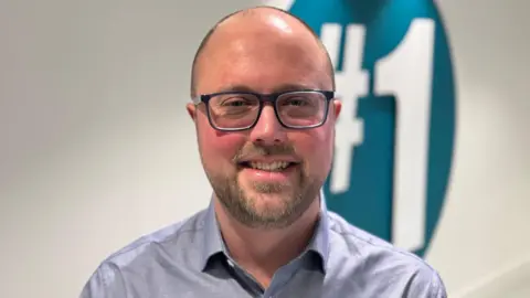 BBC Olly Newton smiling. He is wearing a blue shirt and dark rimmed glasses. He has short hair and a beard.