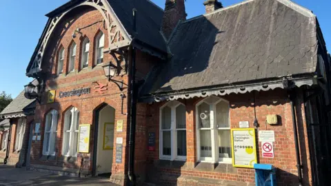 BBC News/Claire Hamilton A red-brick Victorian railway station, with an ornate grey slate roof and large white-framed windows. Above the door is a sign saying Cressington, alongside the insignia of Merseyrail and National Rail. 
