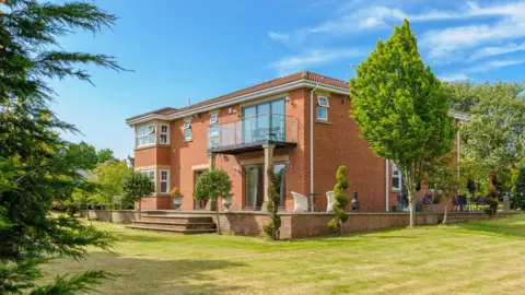 Red Gables. It is a two-storey brown-brick house with a balcony on the second floor and a seating area outside. It is surrounded by trees and other plants.