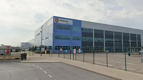 A large warehouse building, viewed from a car park. The corner of the building has blue cladding and an Amazon company logo on a white background at the top.