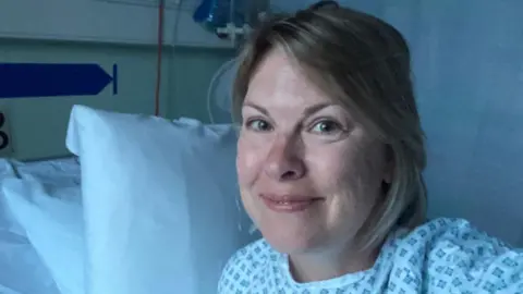 Emma Ward Woman smiles in a hospital gown on a bed in a clinical room with white pillows, curtain and medical equipment.