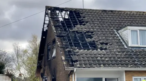 The roof of a bungalow is shown, charred by the fire to the point where there are gaps in the roof. The whole house isn't burned, and some trees can be seen in the background.