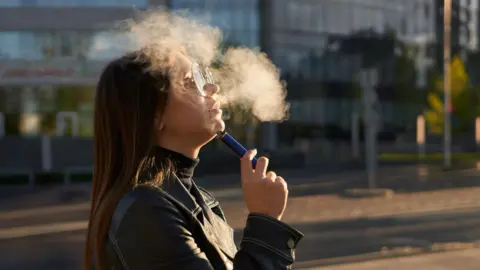 A woman with long brown hair and glasses is smoking a disposable vape. There is a puff of white smoke in the air. She is wearing a black jacket and turtle neck as she stands on the street.