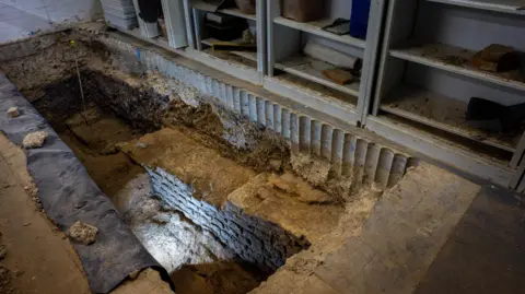 Tony Jolliffe/BBC A section of stone wall about three metres in length is clearly visible at the bottom of a trench dug in the basement of a building next to a row of filing cabinets and shelves. The Roman wall has about seven or eight layers of stones, is about one metre wide and is vey well preserved. 