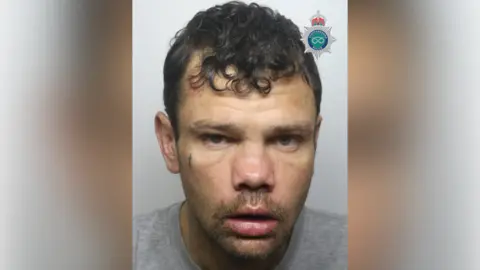 A police mugshot of a man with short black hair, a tiny crucifix tattoo on his right cheek, a short beard and a grey top.