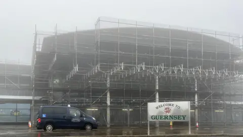 Guernsey airport with fog. A blue taxi is parked at the front. There is also a sign saying welcome to Guernsey. The airport building is covered in scaffolding,