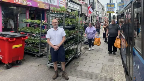 Peter Gallagher a man in shorts, a shirt and boots stood outside his business where there are trolleys of flowers behind him and a St George's Cross flag flying in the distance. On the pavement next to the shop is a bus stop and people can be seen queueing for a bus which has just arrived.