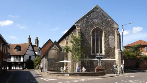 Historic Norfolk An image of Becket's Chapel in Wymondham, Norfolk. The stone building has arched windows and an arched roof. There are properties either side with a road in front.