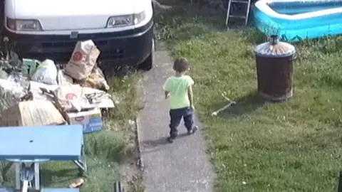Toddler seen from behind walking down garden path in yellow t-shirt and dark trousers. There is a white van and rubbish on the grass as well as a bin and a paddling pool.