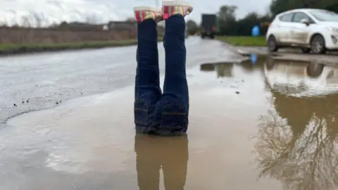 Sophie Montague/BBC A pair of fake legs are placed in a water-filled pothole with the feet sticking out at the top. The legs are constructed from blue jeans and there are colourful shoes on the fake feet. The pothole is large and is at the side of a road. The water is muddy. There is a white car in the background and a green lorry further down the road.