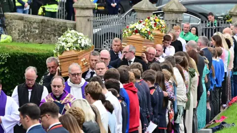 Pacemaker Three light brown coffins with large flower bouquets placed on top are carried up to St Mary's Church in Maguiresbridge. There are four priests in white and purple vestements leading the congregation. On the side of the path, people are stood with their backs to the camera forming a guard of honour as the remains are carried in.