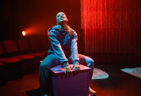 Charlie Flint Emily Glaze sits on a stage on a wooden box, wearing a blue hoodie and jogging bottoms. A red sparkly curtain and red theatre seats are behind her.