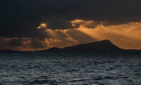 Kate Masson Light shines through dark clouds over a hill and loch