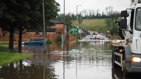 Simon Burns Flooding on the main Enniskillen to Dublin Road in Enniskillen on Sunday evening. Part of the road is submerged in flood water. One blue hatchback car is parked on the left of the photo. The water level is up to its rear bumper and more than half way up its rear wheel arch. A large white truck used to clear drains is in the foreground and cars are turning away from the area in the background.