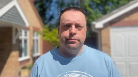 Simon Thake A middle aged man in a blue t-shirt looks dejected towards the camera outside a red brick house