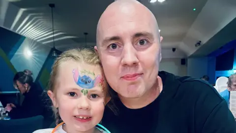 Chris Capstick is sitting next to his daughter Isabella in a restaurant. They are smiling at the camera for this posed photograph. 