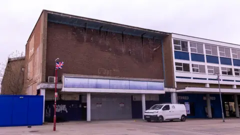 Harlow Council A large brick building that once housed a cinema. A parade of shops has been built next to it, with flats above them.