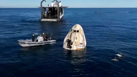 Dolphins are seen swimming next to a domed capsule with a rib motorboat floating next to it and a carrier boat in the background, on a deep blue sea with blue sky in the background, shortly after the astronauts splashed down in the Atlantic off the coast of Florida, on 18 March