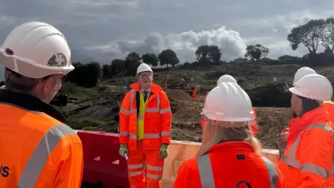 Group of people in high-vis clothing in front of a building site, all with backs to the camera except Richard Clewer who is talking to the group