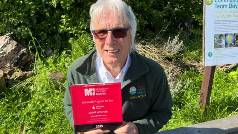 Brian Pearce, 76, has a full head of grey hair. He is wearing dark glasses and a green fleece top with the logo on from Railworld Wildlife Haven. He is holding an award made of glass that is engraved.