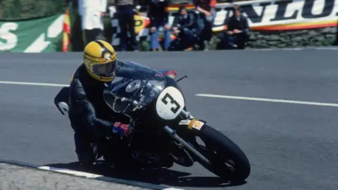 Getty Images Joey Dunlop racing one of his bikes at the Isle of Man, it is black and has a white circle with the number 3 on it. He is wearing a black racing suit and a yellow helmet. He is coming around a corner on the bike as the image is taken. There are spectators in the background watching.