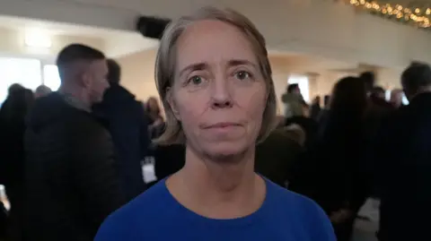 Qays Najm/BBC Sophie Marple standing inside a hall at a meeting. She is looking directly at the camera and is smiling. She is wearing a blue T-shirt.