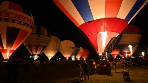 Cameron Balloons Around ten inflated hot air balloons sitting on the grass. It is dark and the balloons are lit up with fire from the inside and a large crowd of people are gathered around to watch.