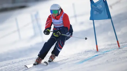 Special Olympics Annabelle Lamb ski ing on a snowy slope, with a blue flag to the right of the image. She is wearing navy trousers with GREAT BRITAIN on the thigh, an orange top, white helmet and blue/green goggles.