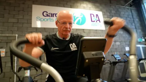 Gordon Weir, a 59-year-old bald man with glasses. He is pictured in the gym of Dundee and Angus College's Gardyne Sports Centre on a cross trainer and is wearing a black college T-shirt.