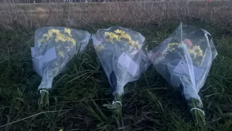 Janine Machin/BBC A close-up of three bunches of flowers placed on a grassy bank next to the river.