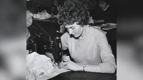 A woman works a sewing machine in a factory. A number of other women are behind her sitting at machines. She is wearing a wristwatch and pearl necklace and a light coloured jumper.