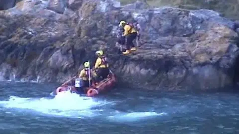An RNLI crew in an orange boat help two children onto the vessel during a rescue operation. Two of the crew are in the boat while another is on the rocky ledge.