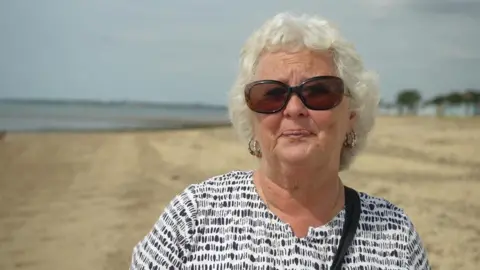 Jamie Niblock/BBC Rita Coombs wears a patterned white top, has black sunglasses on and a white-blonde perm haircut. She is on a beach, facing the camera, pursing her lips.
